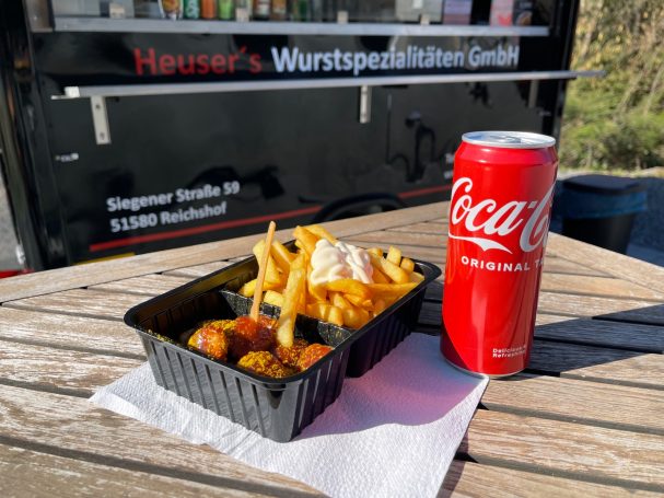 Pommes, Fleischbällchen und eine Coca-Cola-Dose auf einem Tisch vor einem Imbisswagen.